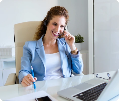 A woman wearing a headset and a light blue blazer smiles while sitting at a desk and looking at her laptop. She holds a pen and appears to be engaged in a video call or virtual meeting with letting Agents